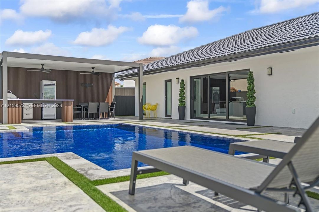 Outdoor living space featuring a covered patio, built-in kitchen station with bar seating, swimming pool, and stone-finish decking with artificial turf accents