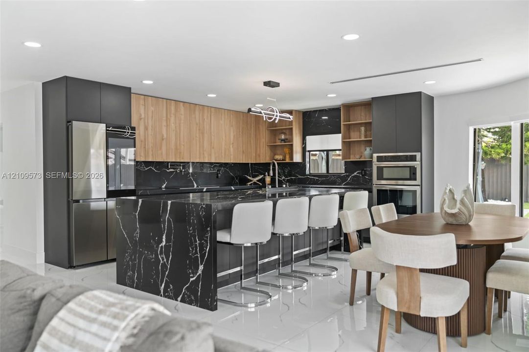 Modern kitchen featuring two-tone cabinetry with wood-finish uppers, a central island with waterfall-edge countertop, dark stone slab backsplash, integrated stainless steel appliances, and recessed lighting