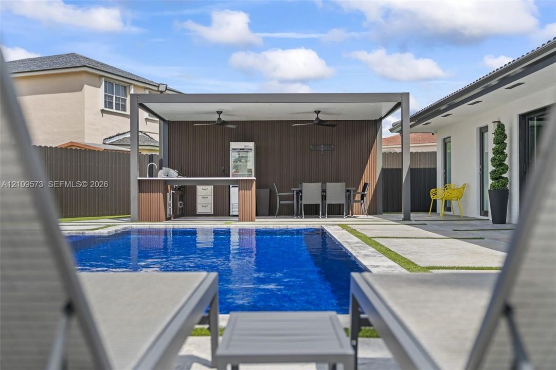 Rectangular swimming pool with stone coping, concrete paver patio with turf inlay, and a covered outdoor kitchen featuring a built-in grill, cabinetry, and ceiling fans
