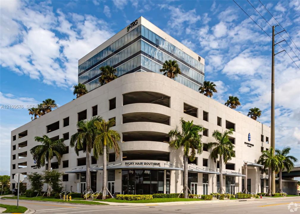 Mixed-use commercial structure featuring a glass-paneled mid-rise office tower, multi-level concrete parking garage, ground-floor retail storefronts, and perimeter palm tree landscaping