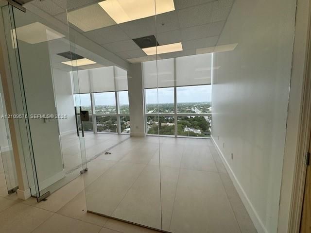 Office space featuring floor-to-ceiling glass wall partitions, ceramic tile flooring, recessed grid ceiling panels, and expansive windows with integrated roller shades