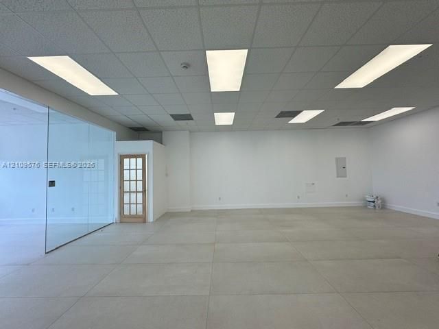Open-concept interior featuring large-format tile flooring, recessed drop-ceiling grid, glass partition wall, and a multi-pane wood-framed door