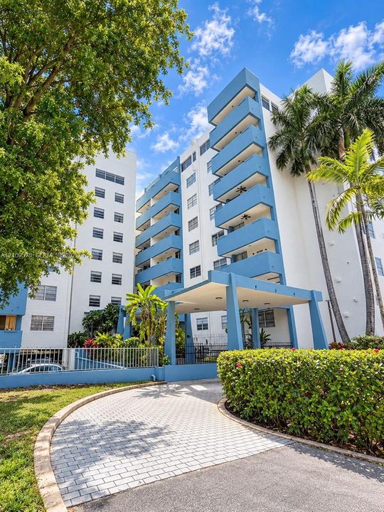 Mid-rise multi-story exterior featuring a covered porte-cochère, stacked private balconies, paved circular driveway, and mature landscaping