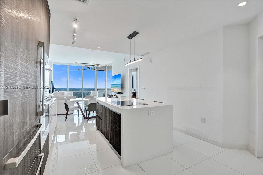 Modern kitchen featuring a central island with a solid-surface countertop and integrated cooktop, textured dark cabinetry, white tile flooring, recessed track lighting, and floor-to-ceiling windows