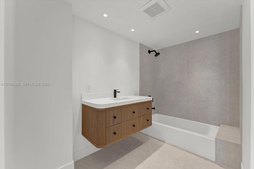 Floating wood-finish vanity with a white countertop and rectangular undermount sink, matte black faucet and shower fixtures, built-in bathtub with a stone-finish tiled surround, and recessed ceiling lighting