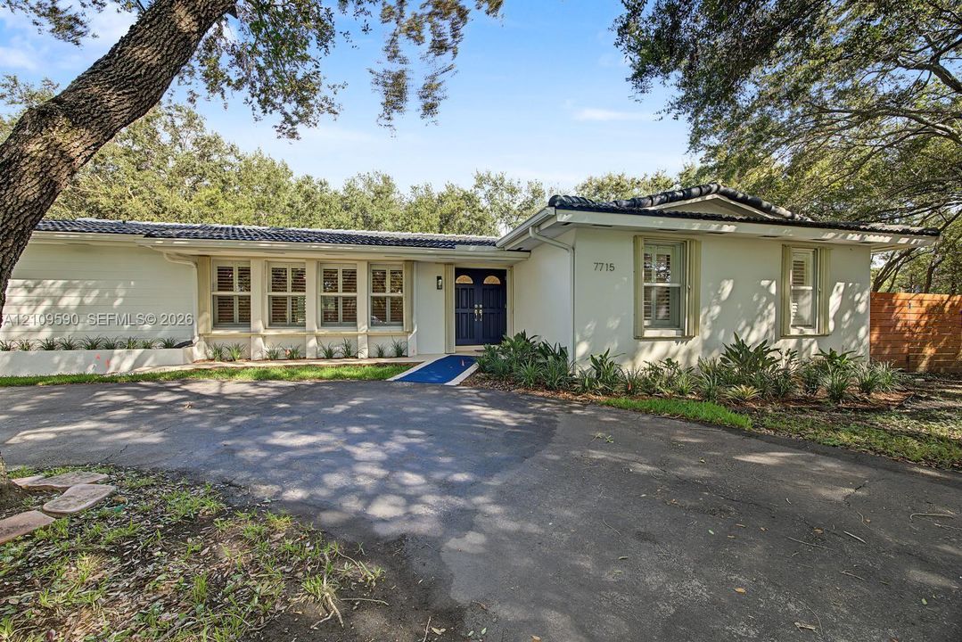 Single-story exterior featuring a light-toned masonry facade, dark tile roof, double-door entryway, and paved circular driveway surrounded by mature trees
