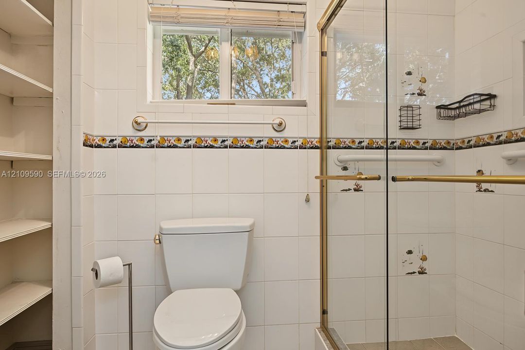 Bathroom featuring white wall tiling with decorative accent trim, glass-enclosed shower with gold-tone hardware, built-in recessed shelving, and a double-pane window