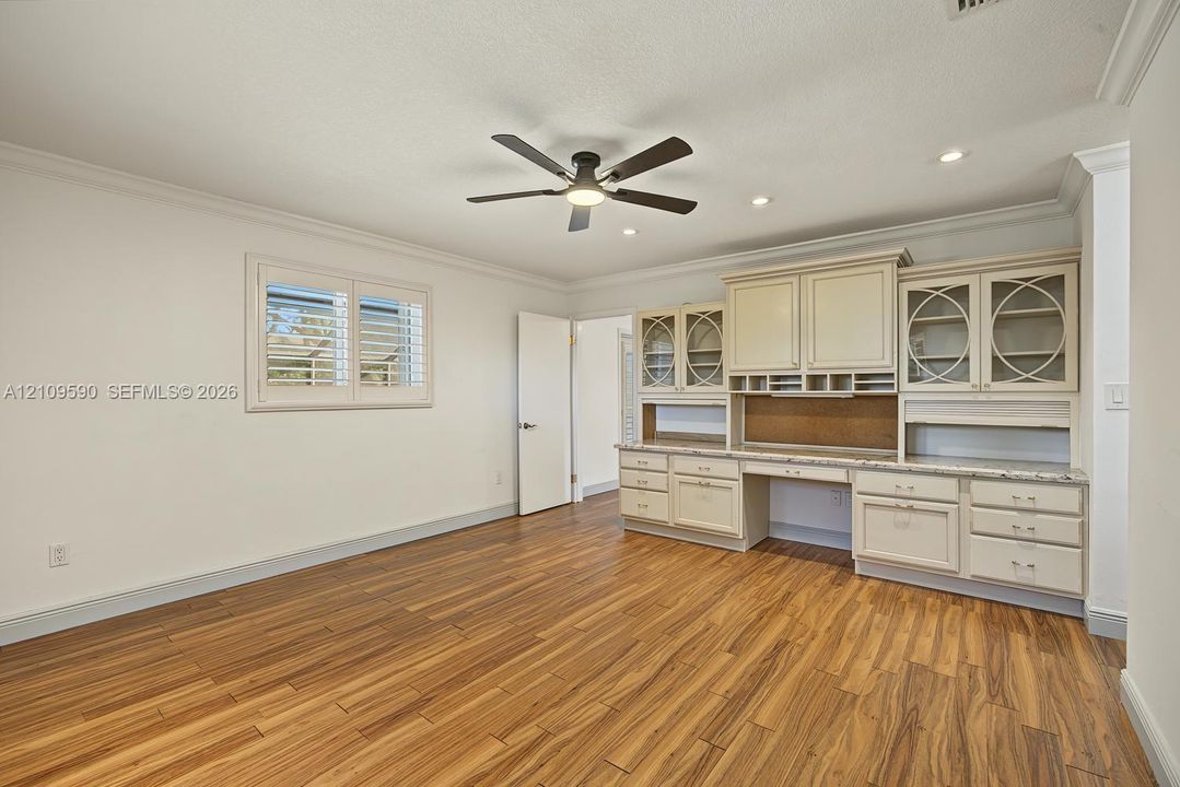 Wood-finish flooring, built-in desk with overhead cabinetry, ceiling fan, and plantation shutters