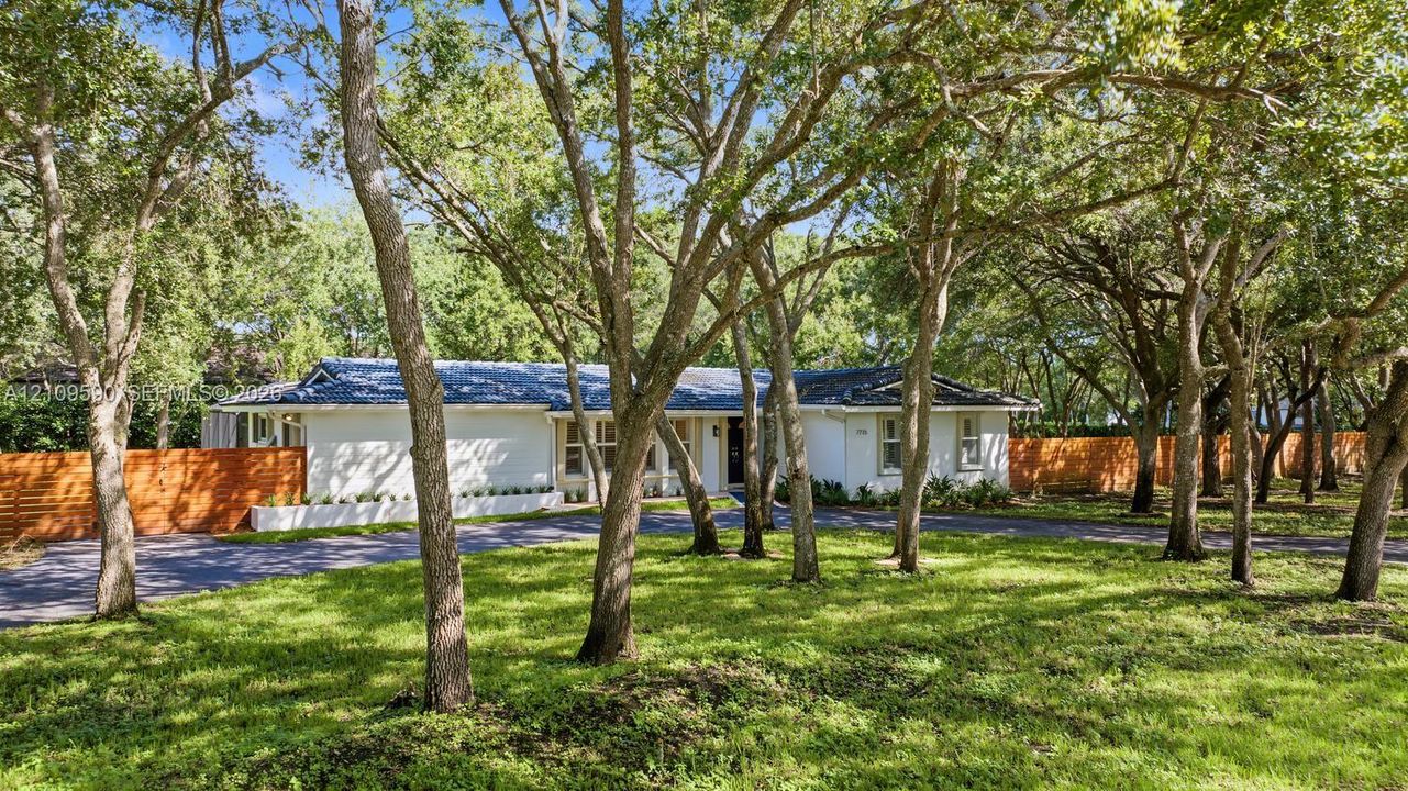 Single-story exterior featuring painted masonry siding, a dark-toned shingled roof, horizontal wood-slat fencing, and a paved driveway, all set within a landscape of mature trees