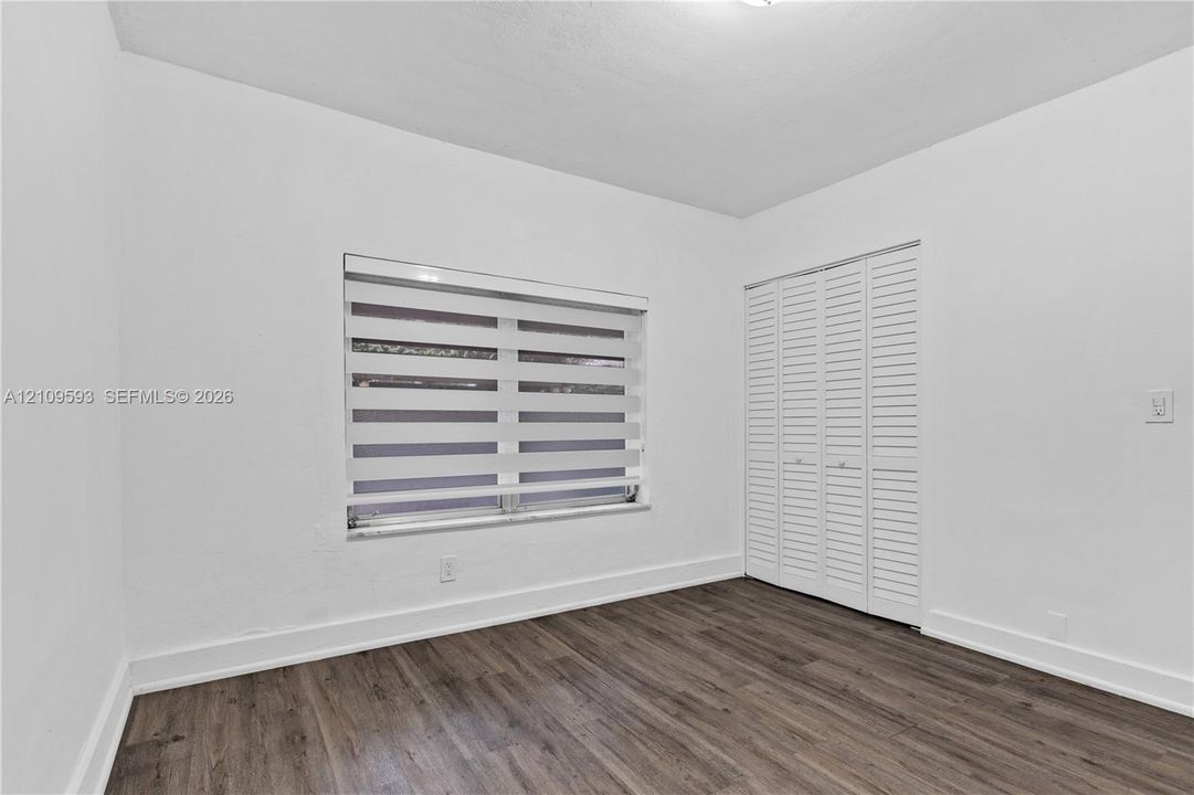 Room interior featuring wood-finish flooring, white walls, louvered bi-fold closet doors, and a window with horizontal dual-shade blinds