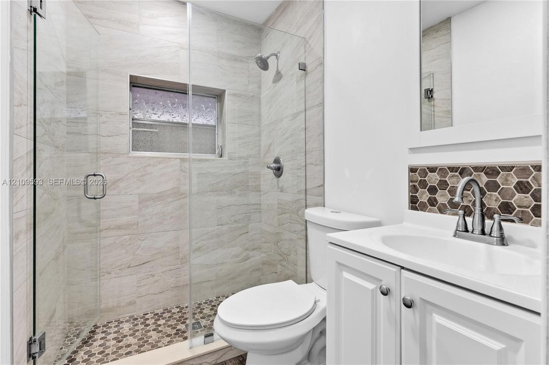 Walk-in shower with frameless glass enclosure, floor-to-ceiling neutral tile surround, pebble-style shower flooring, white vanity with integrated sink, and honeycomb tile backsplash