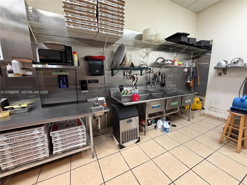 Commercial kitchen space featuring stainless steel wall panels, a triple-basin industrial sink, wall-mounted wire shelving, an integrated ice machine, and tiled flooring