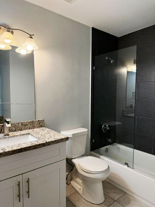 White vanity with stone-finish countertop, undermount sink, three-light fixture, bathtub with glass panel, and dark tile surround