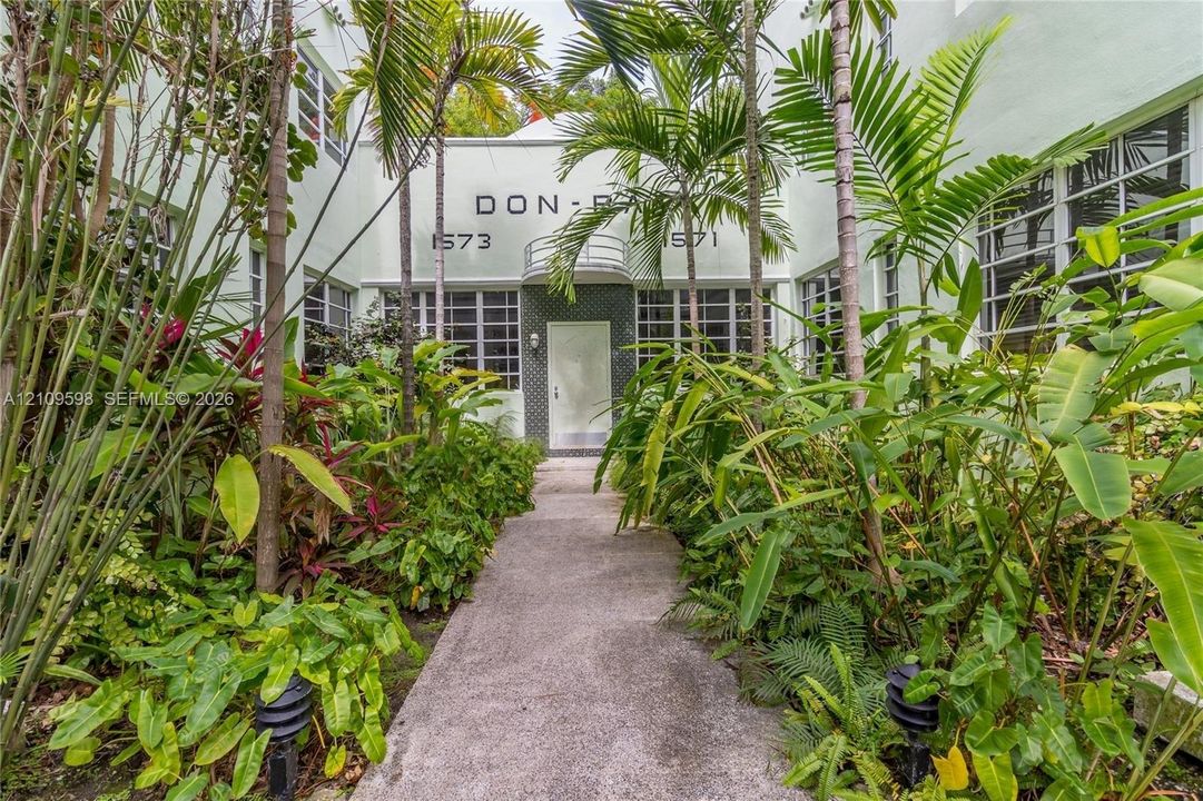 Art Deco exterior featuring a concrete walkway, mature palm trees, multi-pane windows, and a central entry with a rounded metal railing
