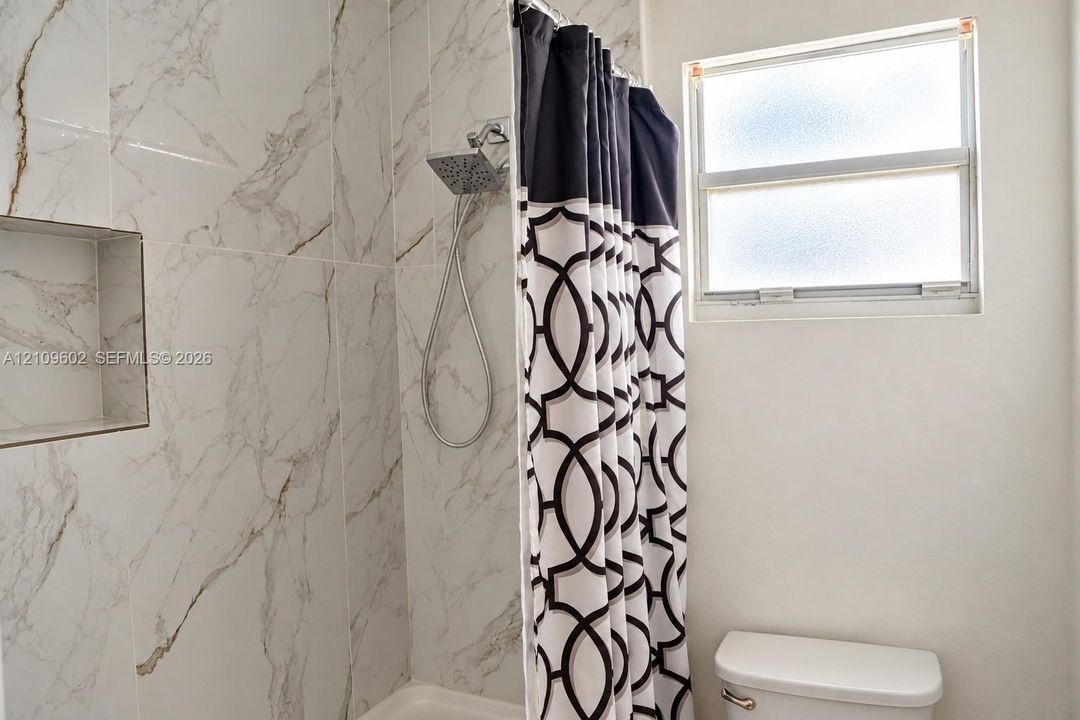 Shower area featuring full-height stone-finish tile surround, integrated recessed wall niche, rain-style showerhead with handheld attachment, and a single-hung frosted glass window