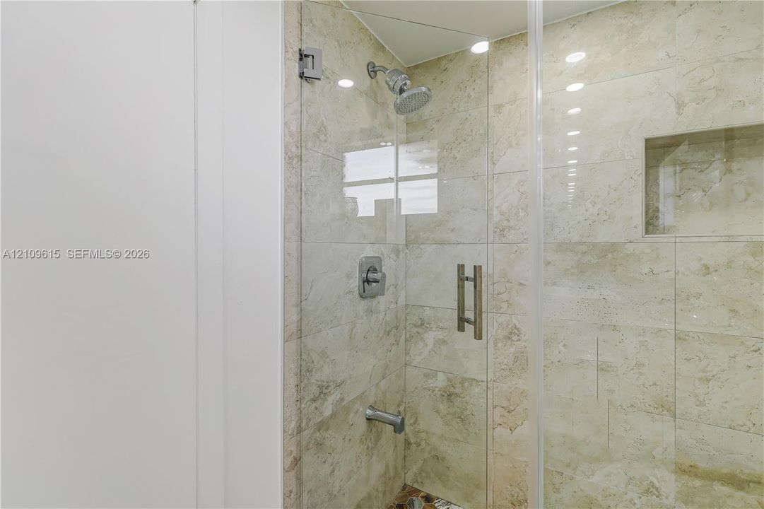 Walk-in shower featuring floor-to-ceiling stone-finish wall tile, frameless glass door with stainless steel hardware, integrated recessed storage niche, and brushed nickel fixtures