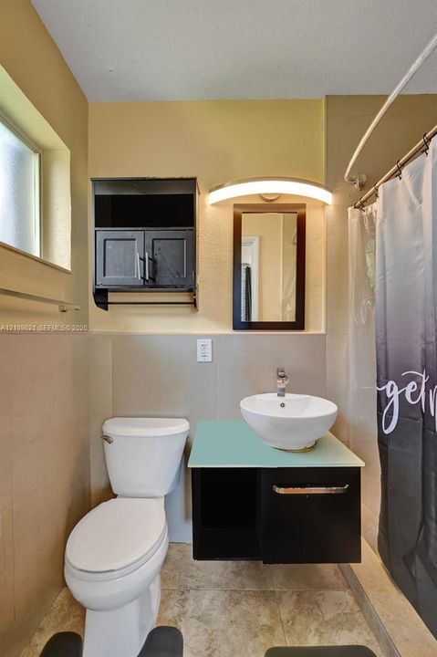 Wall-mounted floating vanity with a frosted glass countertop and white vessel sink, complemented by matching dark cabinetry, a wall-mounted arched light fixture, and stone-finish floor tiling