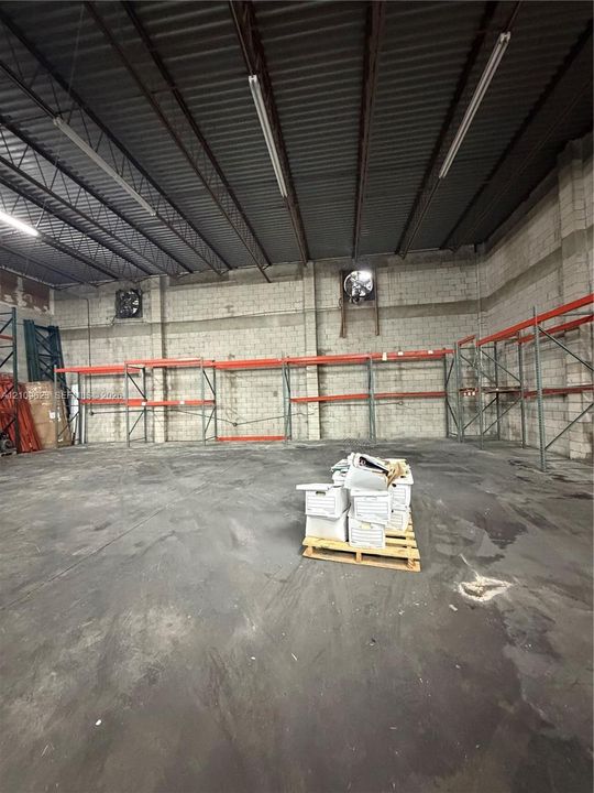 Industrial warehouse space featuring high ceilings with exposed metal trusses, concrete masonry unit walls, integrated wall-mounted exhaust fans, concrete flooring, and built-in metal racking systems