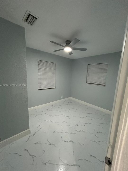 Neutral-toned room featuring large-format tile flooring with marble-patterned veining, dual windows with fitted blinds, and a multi-blade ceiling fan with integrated lighting