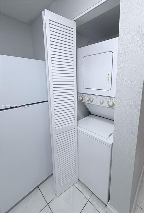 Stacked laundry unit behind louvered door, white refrigerator, and white tile flooring