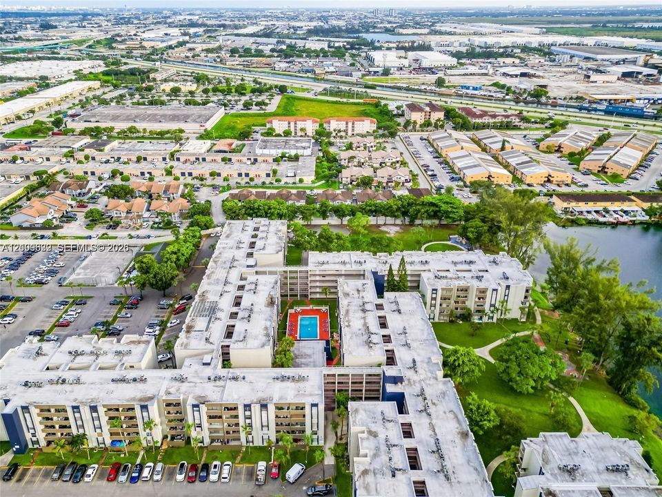 Aerial perspective of a multi-story residential complex featuring a central swimming pool, paved surface parking, manicured green space, and proximity to a canal