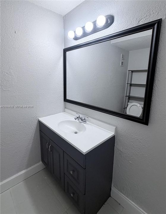 Dark-toned vanity with shaker-style cabinetry, white integrated countertop and sink, polished chrome faucet, rectangular wall mirror with a black frame, and four-bulb vanity light fixture