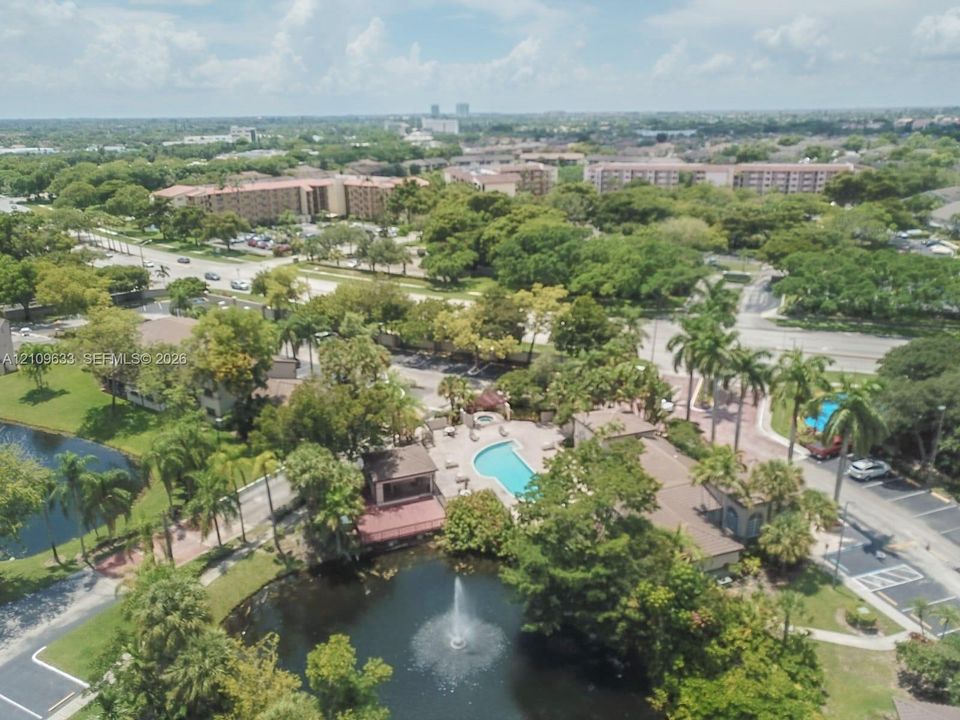 Aerial perspective featuring a swimming pool, a pond with an integrated fountain, mature palm trees, and paved parking areas