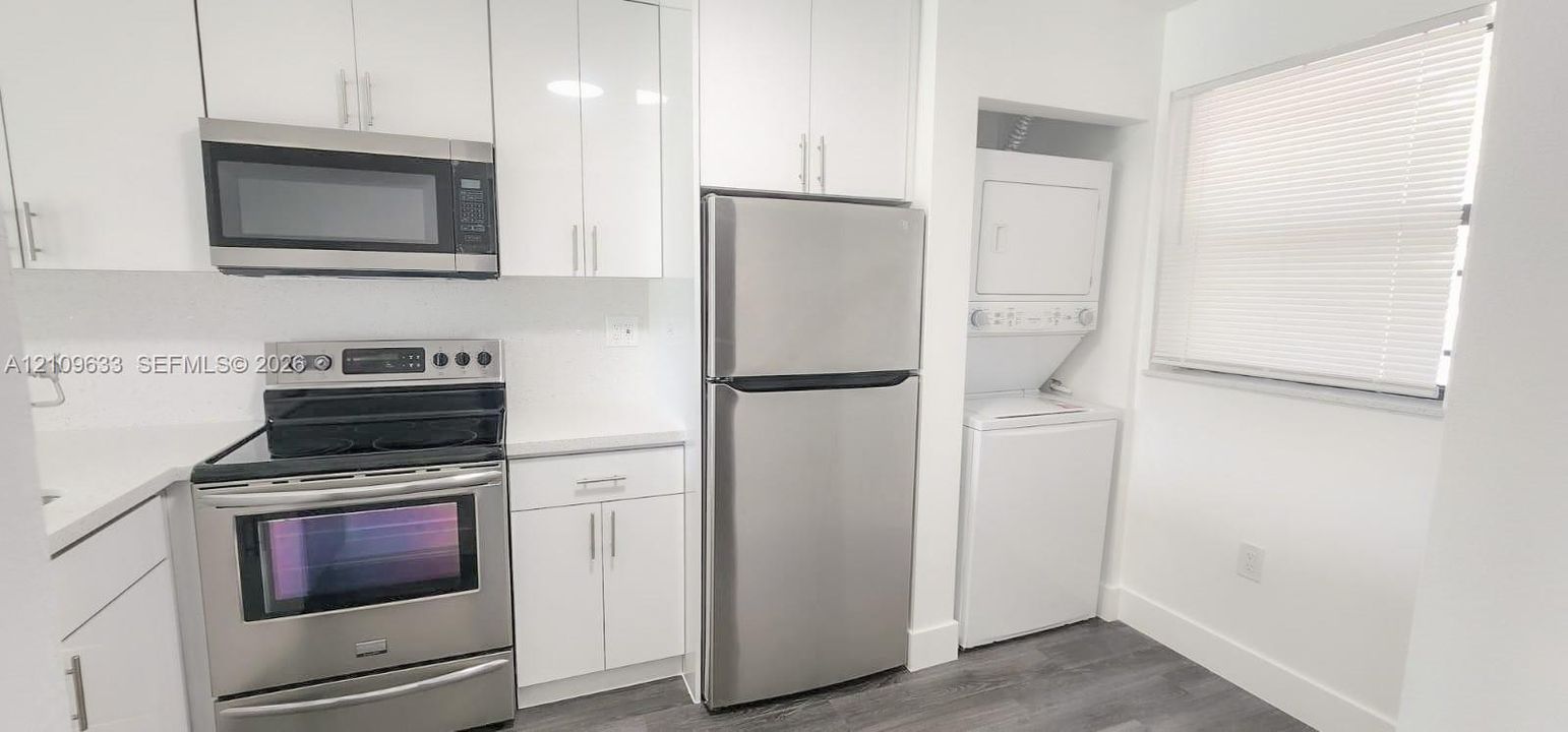 Kitchen area featuring white cabinetry, stainless steel appliances, solid surface countertops, stacked laundry unit, and wood-finish flooring