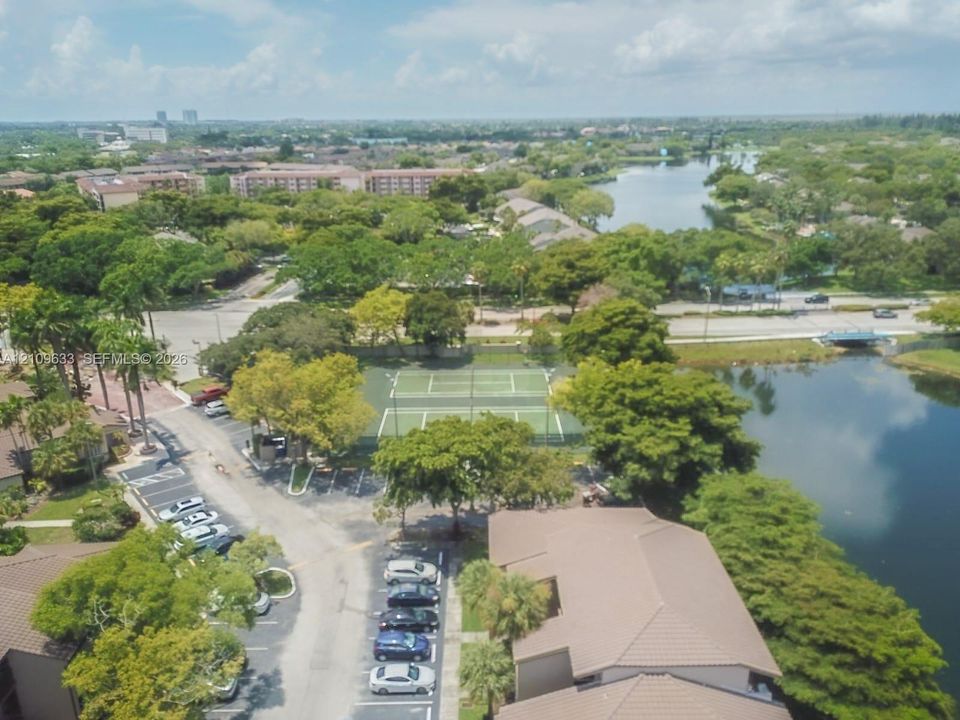 Aerial perspective of a community featuring twin tennis courts, paved parking areas, and mature landscaping situated along a waterway