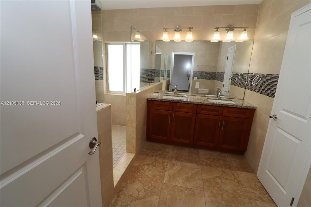 Double vanity with stone-finish countertop, under-mount sinks, wood-finish cabinetry, walk-in shower with glass enclosure, and decorative tile accent wall