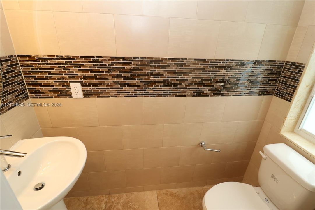 Pedestal sink with chrome faucet, porcelain tile flooring, and wall surfaces featuring large-format beige tiles accented by a horizontal glass mosaic border