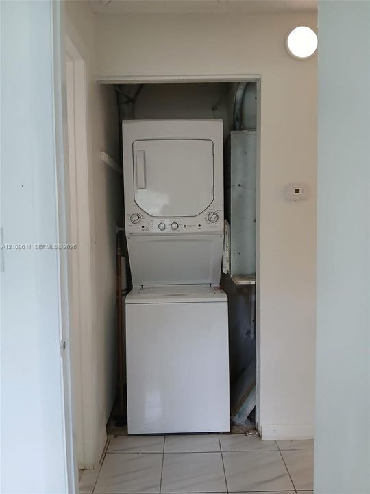 Laundry closet containing a stacked washer and dryer unit, featuring white ceramic tile flooring and a wall-mounted light fixture