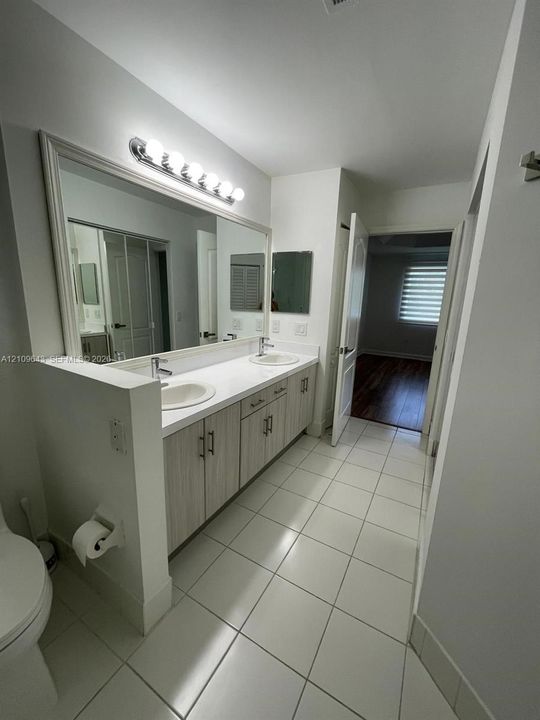 Double vanity featuring light-toned cabinetry, white countertop with dual drop-in sinks, full-width wall mirror, and large-format tile flooring