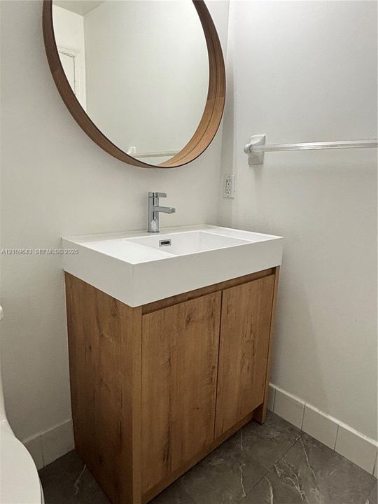 Vanity featuring a wood-finish cabinet, integrated white countertop with rectangular basin, chrome single-handle faucet, and circular wall-mounted mirror with a wood frame