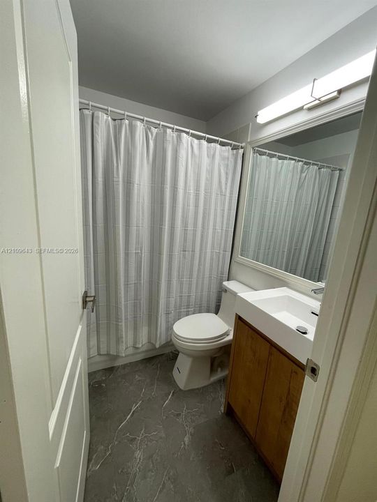 Bathroom featuring a wood-finish vanity with a white rectangular sink, stone-finish flooring, a full-width mirror, and a wall-mounted light fixture