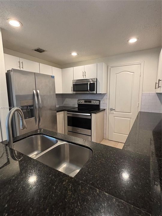 Kitchen featuring dark stone countertops, stainless steel appliances, white subway tile backsplash, white upper cabinetry, and a dual-basin stainless steel sink with a high-arch faucet