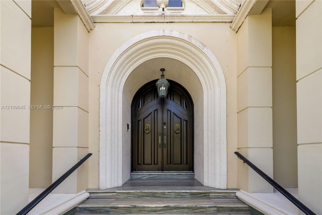 Double arched solid wood doors featuring brass lion-head knockers, set within a recessed masonry entryway with decorative molding, stone-stepped risers, and mounted pendant lighting