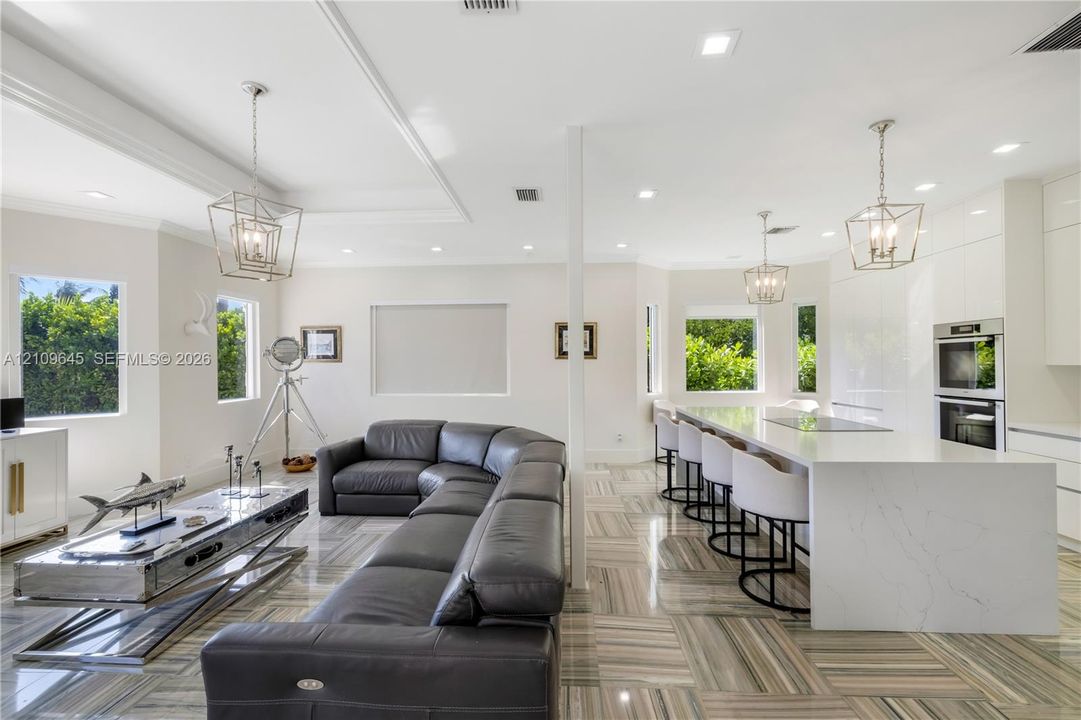 Open-concept interior featuring large-format tile flooring, a waterfall-edge stone island with integrated cooktop, dual built-in wall ovens, recessed lighting, and geometric metal pendant fixtures