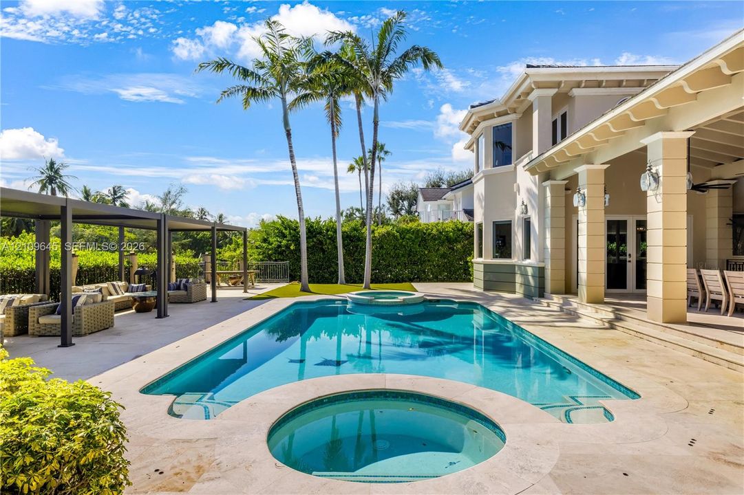 Outdoor pool with an integrated spa, stone-paver pool deck, covered patio with structural columns, and a detached metal-frame pergola