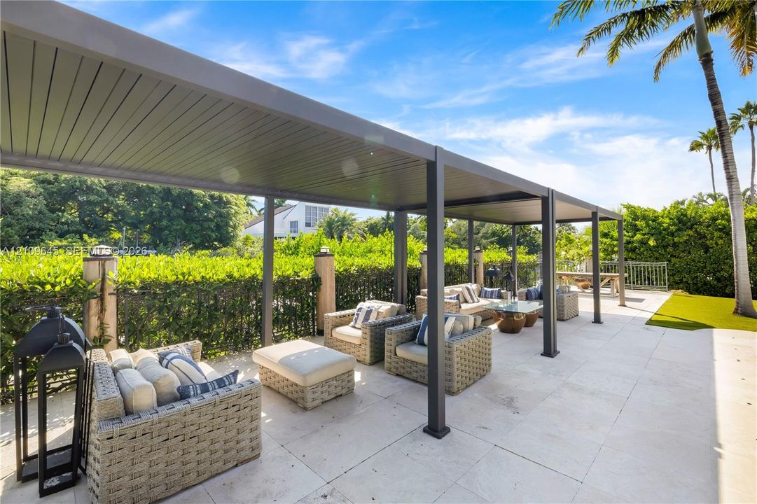 Covered patio featuring a structured metal pergola, large-format stone pavers, and a perimeter of mature privacy hedging
