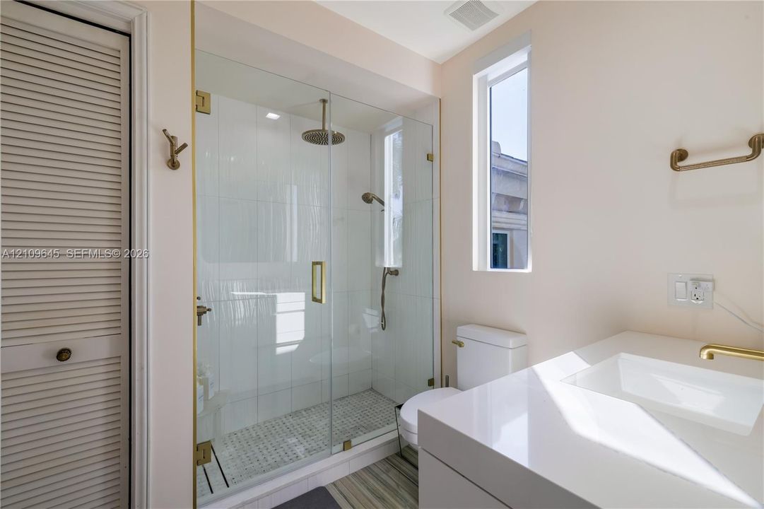 Walk-in glass shower featuring a rain-style showerhead and handheld sprayer with brass hardware, complemented by a white vanity with a rectangular integrated sink and a louvered-door closet