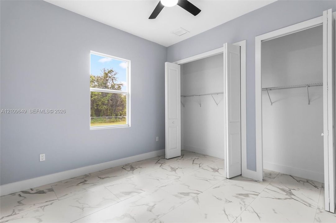 Room featuring large-format tile flooring with a neutral veining pattern, dual closets with bi-fold doors, a single-hung window, and a ceiling fan