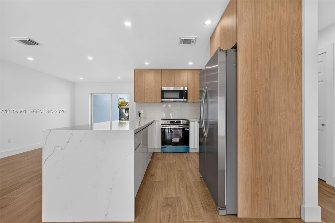 Kitchen featuring an expansive waterfall-edge island, light wood-finish cabinetry, stainless steel appliances, and wood-finish flooring