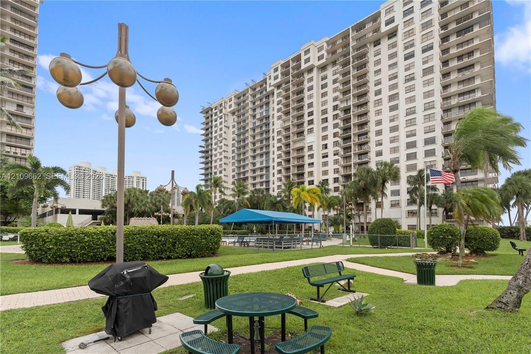 High-rise residential exterior featuring a landscaped lawn with manicured hedges, paved walking paths, fixed outdoor seating, and a covered patio area