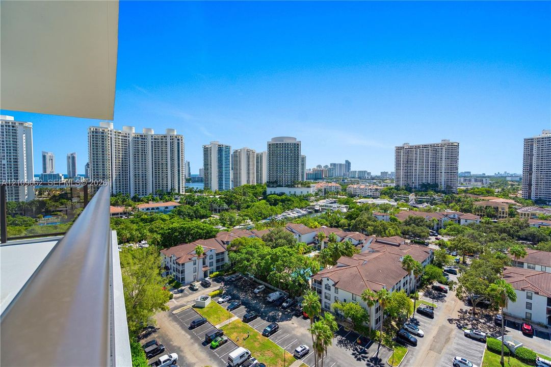 Covered balcony featuring a metal railing and expansive urban surroundings including mid-rise residential structures, mature landscaping, and paved parking areas