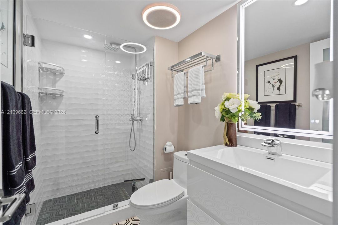 Walk-in shower with a glass enclosure, textured wall tile, and geometric floor tile; wall-mounted vanity with an integrated basin; LED-backlit vanity mirror; and contemporary flush-mount ceiling lighting