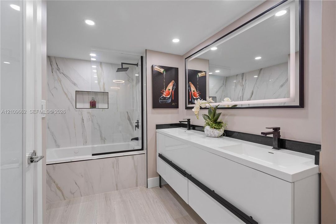 Double vanity featuring white integrated sinks, matte black fixtures, and a backlit mirror, complemented by a soaking tub with stone-finish tile surround, a glass shower panel, and a built-in recessed niche
