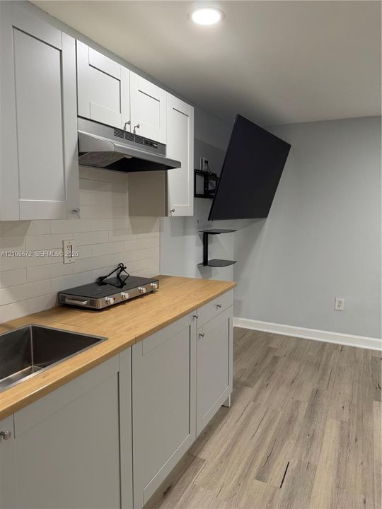 Grey shaker-style cabinetry with wood-finish countertop, stainless steel undermount sink, white subway tile backsplash, and dual floating shelves