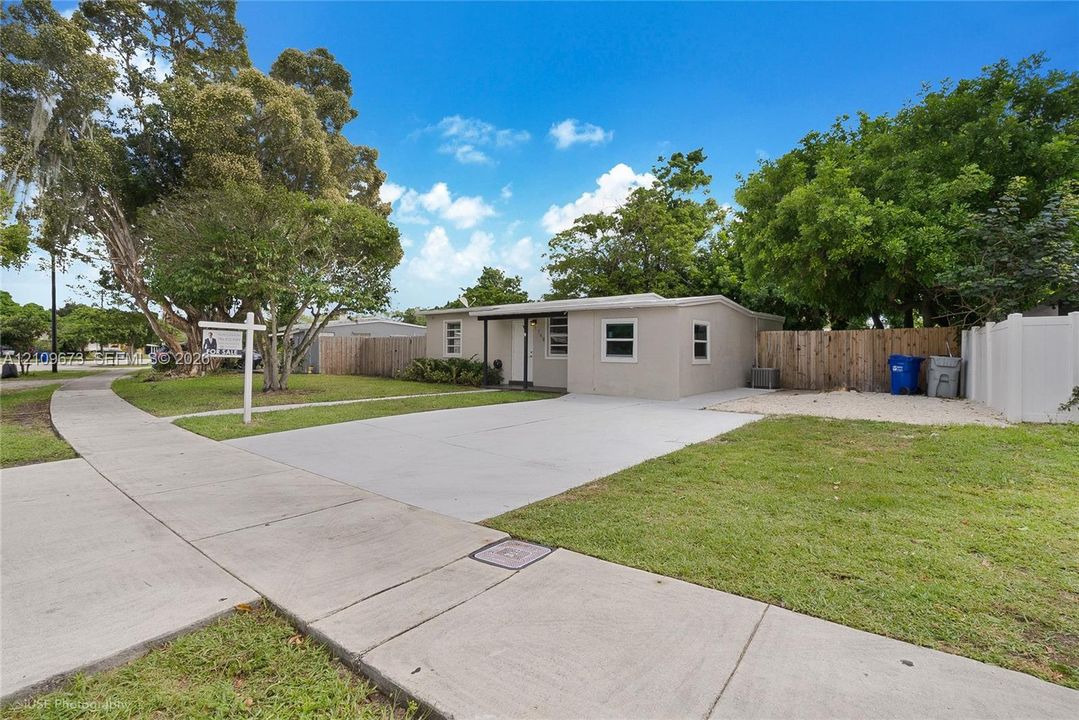 Single-story exterior featuring a concrete driveway, lawn, mature trees, and wood-panel perimeter fencing