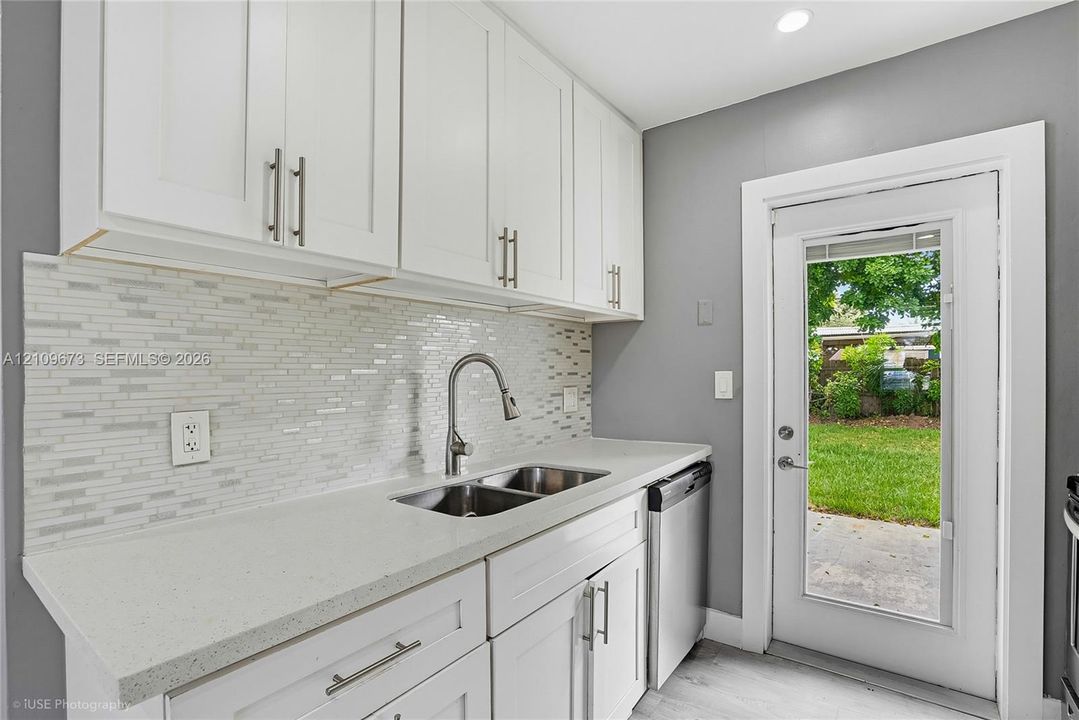 Kitchen featuring white shaker-style cabinetry, solid-surface countertops, horizontal glass tile backsplash, dual-basin stainless steel sink with high-arc faucet, and an exterior glass-panel door